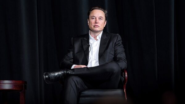 Elon Musk sits in a chair against a dark background