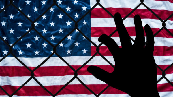 A hand grabs at a chain link fence with an American flag on the other side, symbolizing the struggle to immigrate to America.