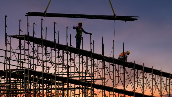 A factory under construction, with a beam being lowered and a crew member standing ready.