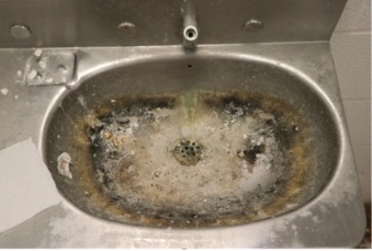 A metal jail cell sink with a water fountain on top; the sink bowl appears severely corroded, with discoloration throughout.